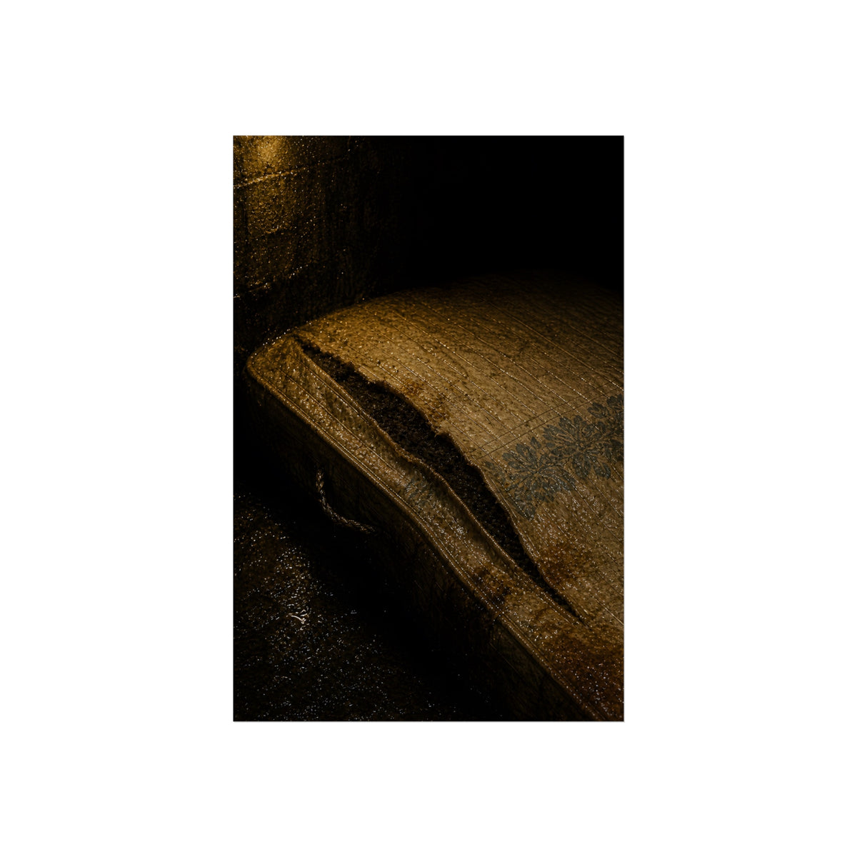 A mattress with its seam split open, lit warm against a dark wall. The Opening of the Ticking by Garabaggio, fine art print, 24″ x 36″.
