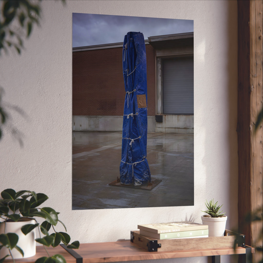 A tall blue tarp bundle roped upright on a wet lot by a loading dock. Folded Standard at the Loading Bay by Garabaggio, fine art print hanging in a room.