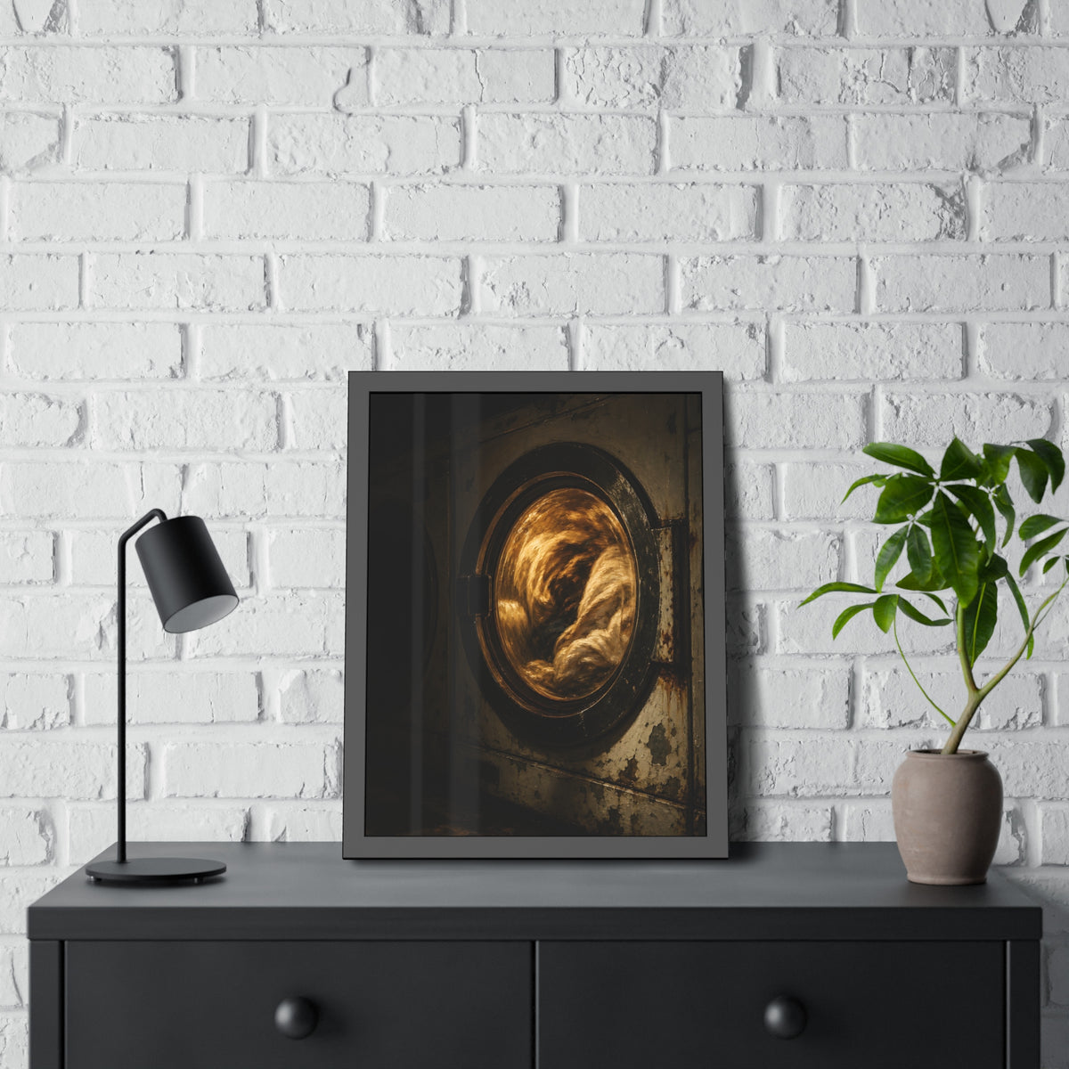 A washer drum tumbling laundry lit amber behind its glass door. The First Spin by Garabaggio, framed fine art print hanging in a room.