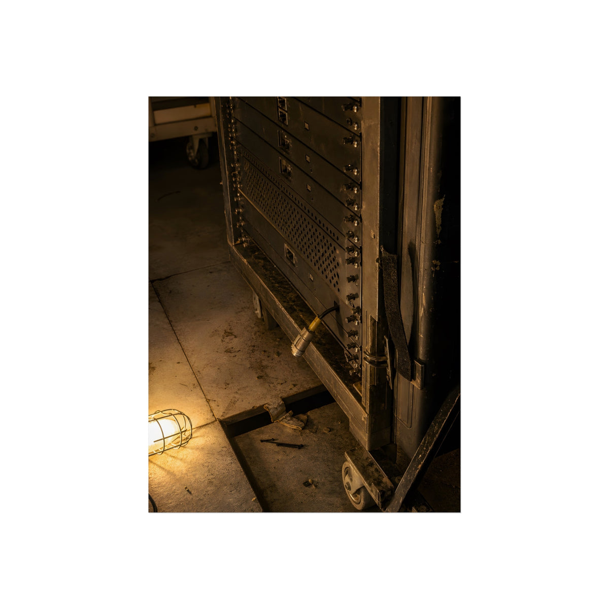 A server rack on a concrete floor beside a caged work light. The Silence at Cutover by Garabaggio, fine art print, 12″ x 16″.
