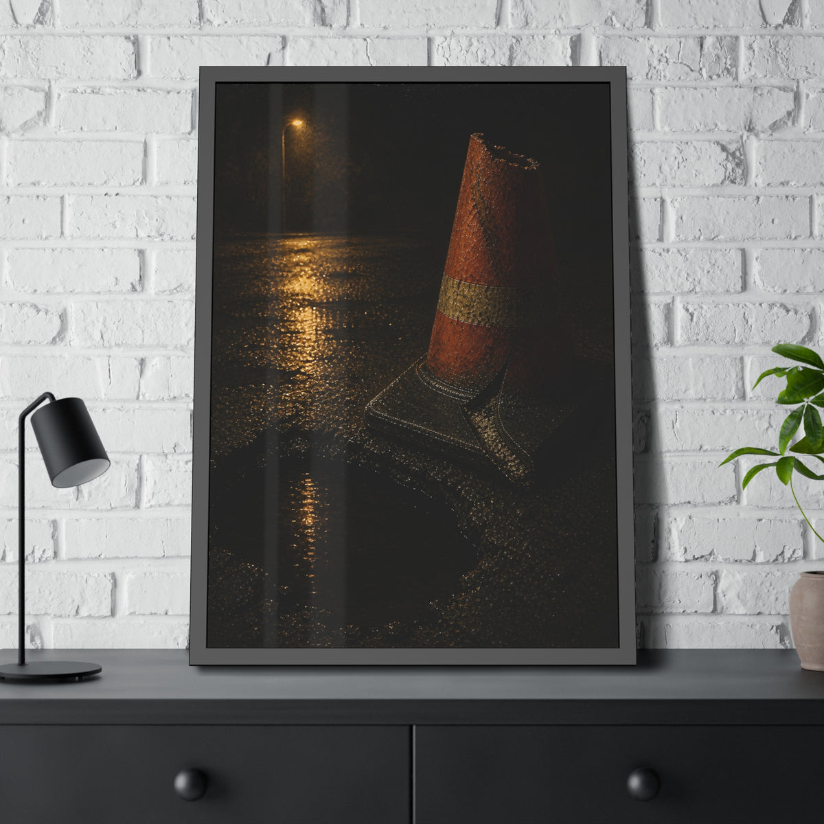 A traffic cone beside a water filled pothole reflecting a street lamp. The Sentinel of the Pothole by Garabaggio, framed fine art print hanging in a room.