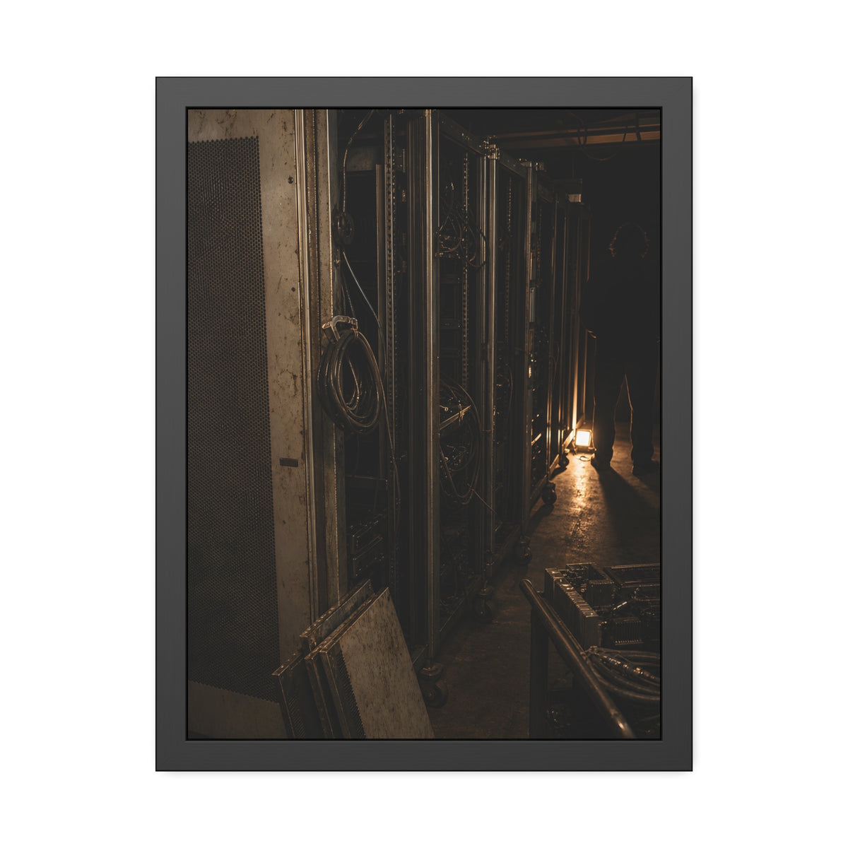 Open server racks in a dark room, a work lamp and a figure at the end. Garabaggio at the End of the Row by Garabaggio, framed fine art print, 18″ x 24″.