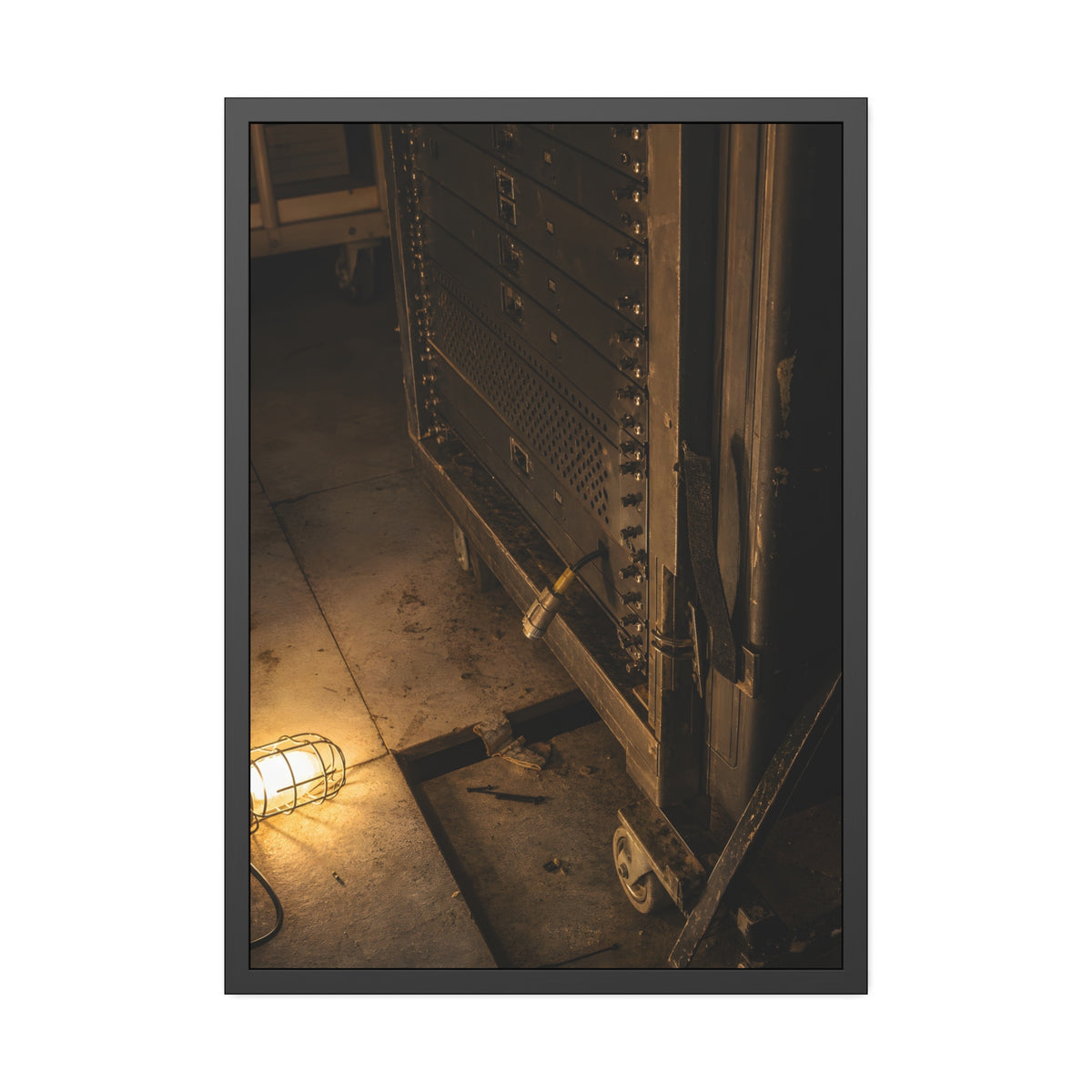 A server rack on a concrete floor beside a caged work light. The Silence at Cutover by Garabaggio, framed fine art print, 28″ x 40″.
