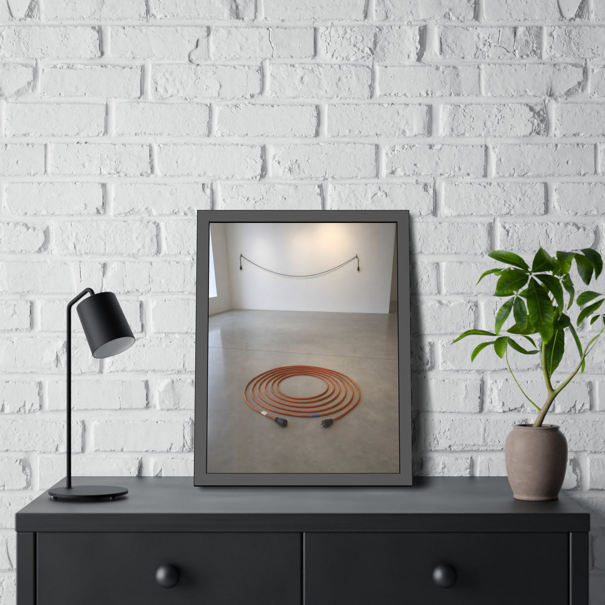An orange extension cord coiled in a spiral on a bare gallery floor. The Floor Coil by Garabaggio, framed fine art print hanging in a room.