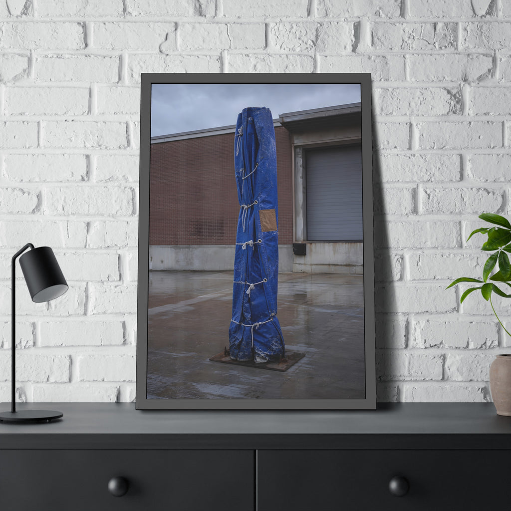 A tall blue tarp bundle roped upright on a wet lot by a loading dock. Folded Standard at the Loading Bay by Garabaggio, framed fine art print hanging in a room.