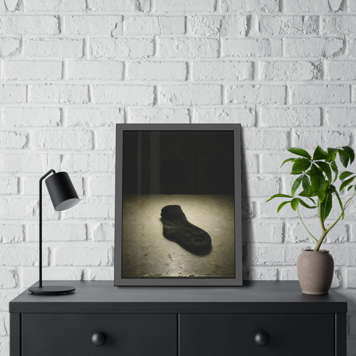 A single dark sock on a laundromat floor before dim washing machines. The Orphaned Sock by Garabaggio, framed fine art print hanging in a room.