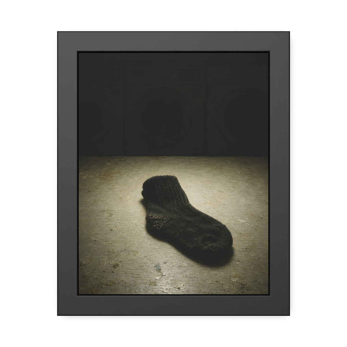 A single dark sock on a laundromat floor before dim washing machines. The Orphaned Sock by Garabaggio, framed fine art print, 11″ x 14″.