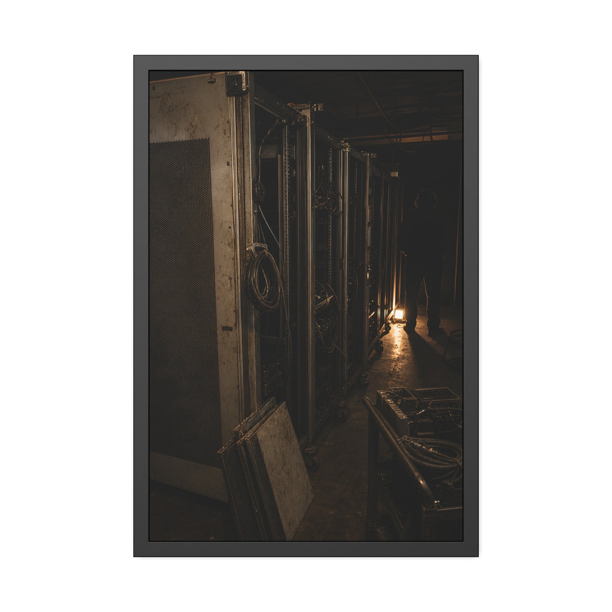 Open server racks in a dark room, a work lamp and a figure at the end. Garabaggio at the End of the Row by Garabaggio, framed fine art print, 24″ x 36″.