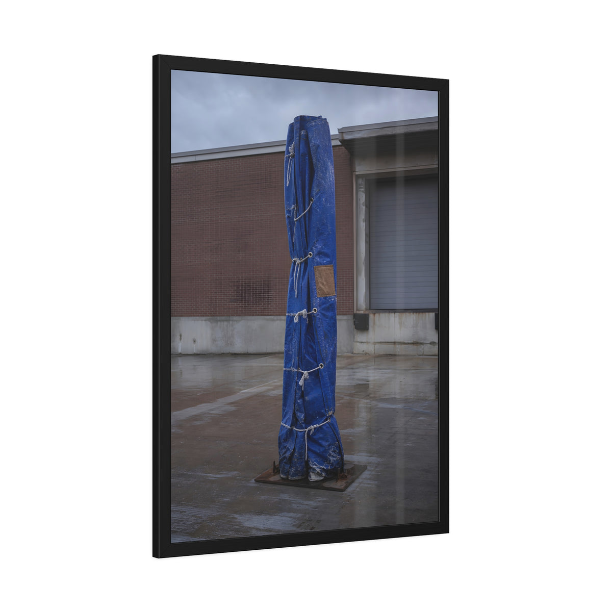 A tall blue tarp bundle roped upright on a wet lot by a loading dock. Folded Standard at the Loading Bay by Garabaggio, framed fine art print hanging in a room.