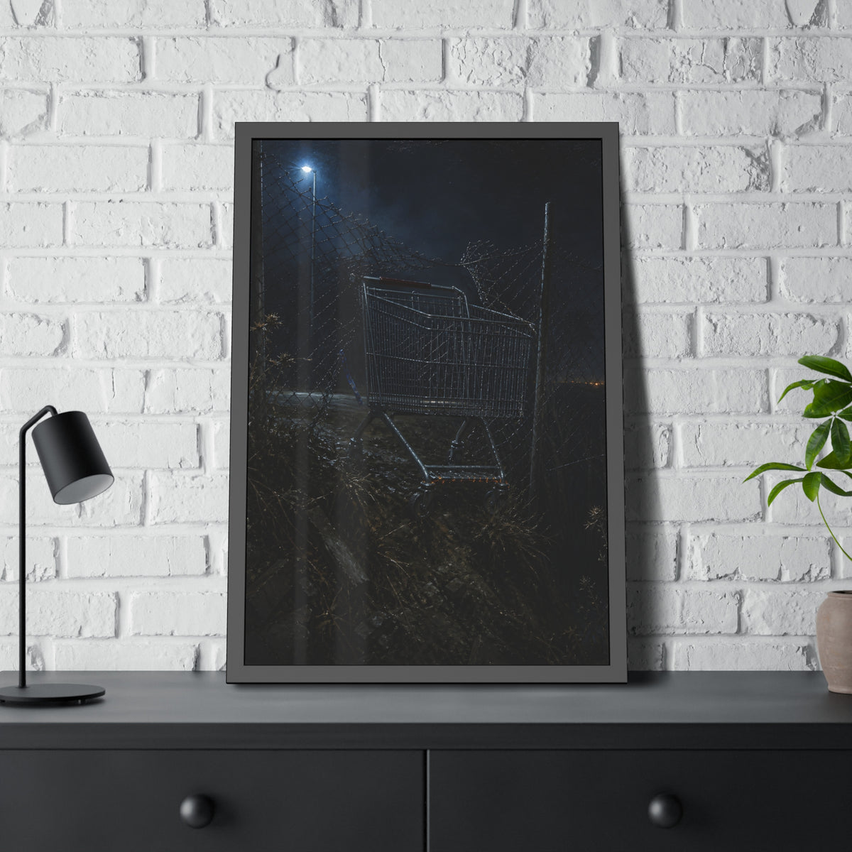 A shopping cart wedged in a chain link fence under a streetlamp. The Abandonment at the Ravine Fence by Garabaggio, framed fine art print hanging in a room.