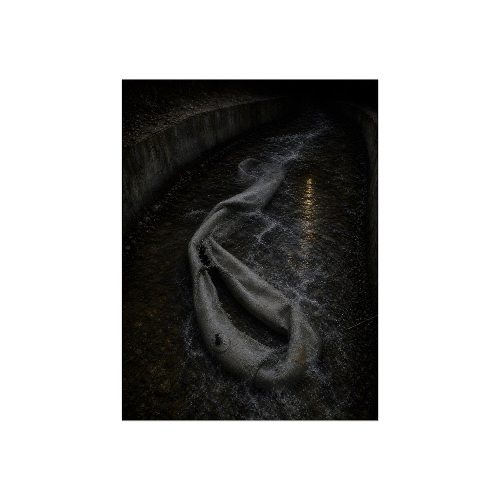 A deflated pool float caught in a concrete drainage channel of running water. The Release into Runoff by Garabaggio, fine art print, 12″ x 16″.