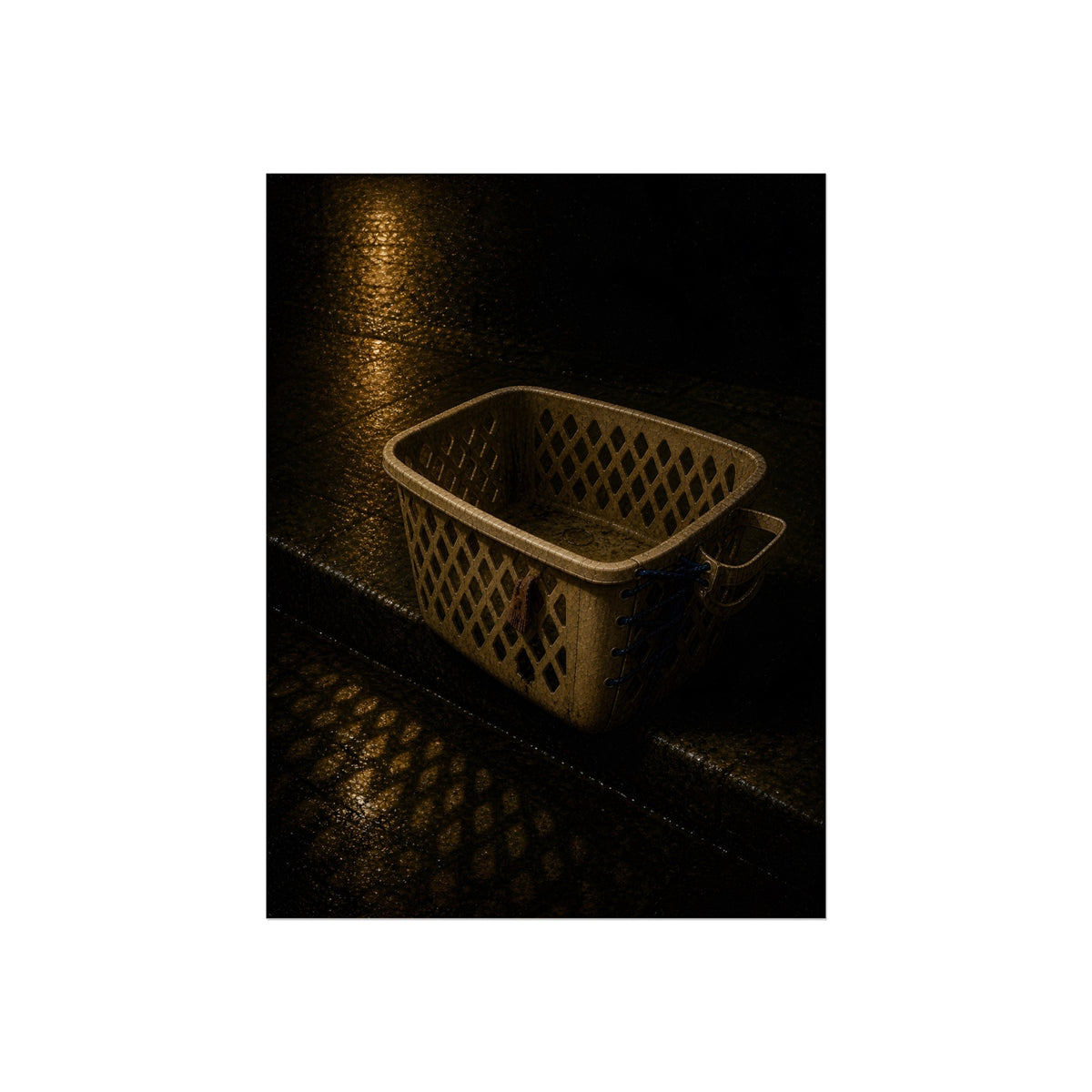 An empty plastic laundry basket on a wet curb under warm street light. The Emptying at the Curb by Garabaggio, fine art print, 12″ x 16″.