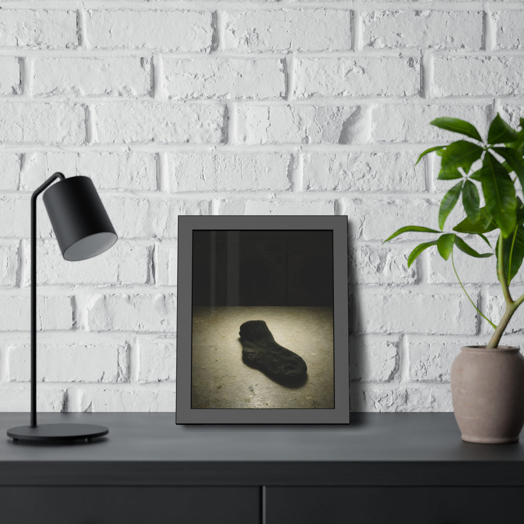 A single dark sock on a laundromat floor before dim washing machines. The Orphaned Sock by Garabaggio, framed fine art print hanging in a room.