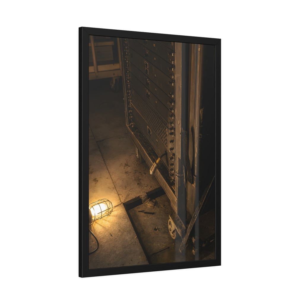 A server rack on a concrete floor beside a caged work light. The Silence at Cutover by Garabaggio, framed fine art print hanging in a room.