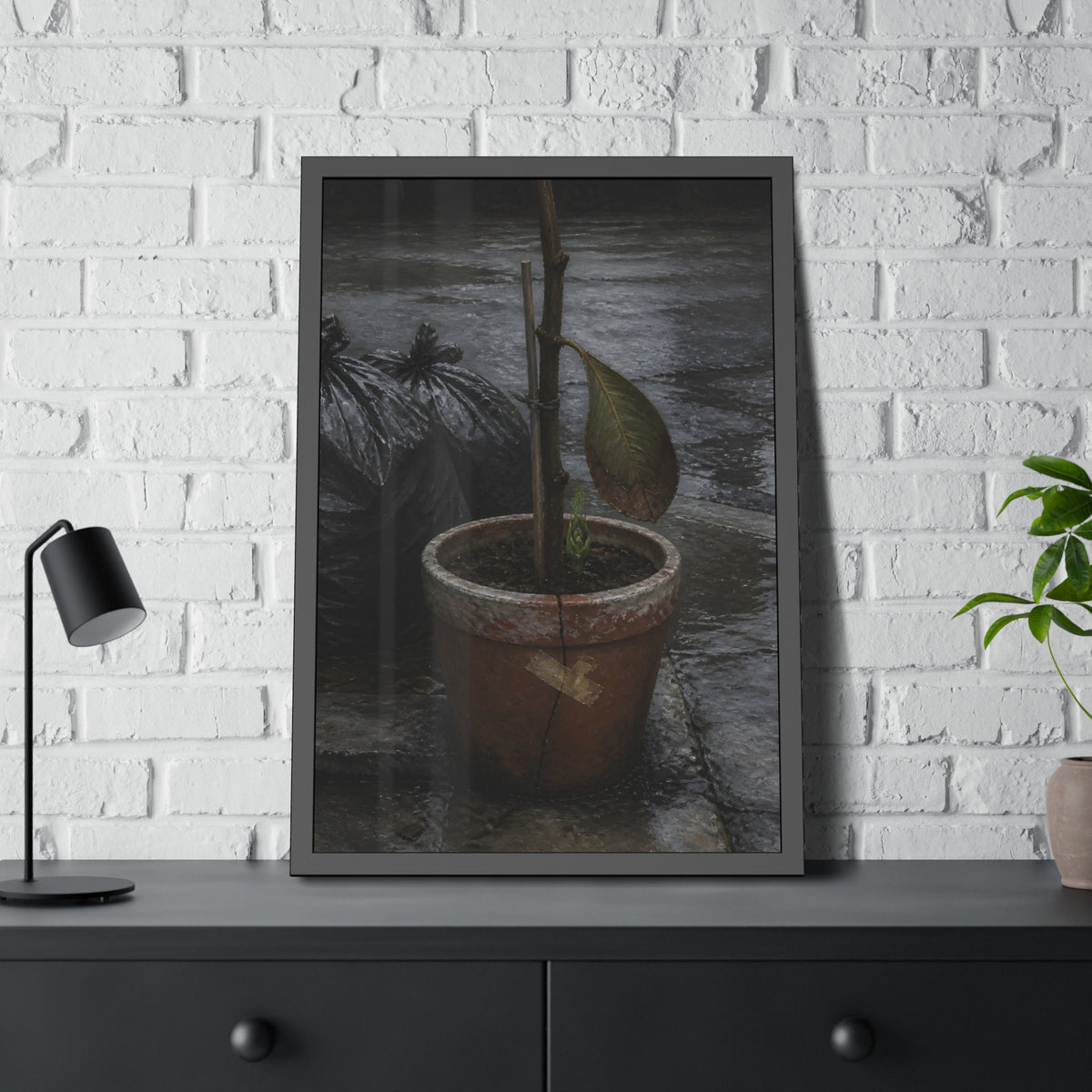 A taped clay pot with one leaf and a green shoot on wet pavement. The Green at the Base by Garabaggio, framed fine art print hanging in a room.