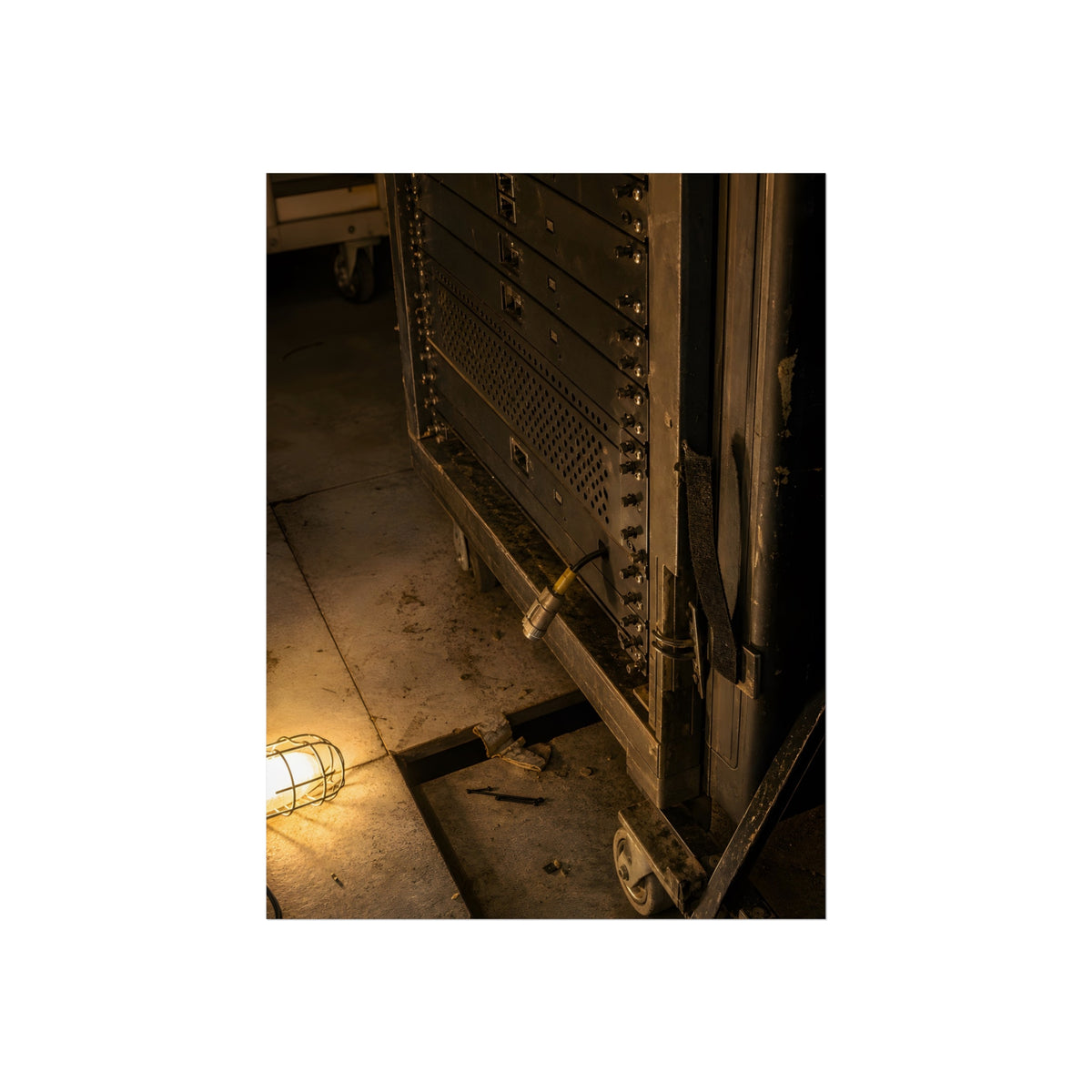 A server rack on a concrete floor beside a caged work light. The Silence at Cutover by Garabaggio, fine art print, 18″ x 24″.