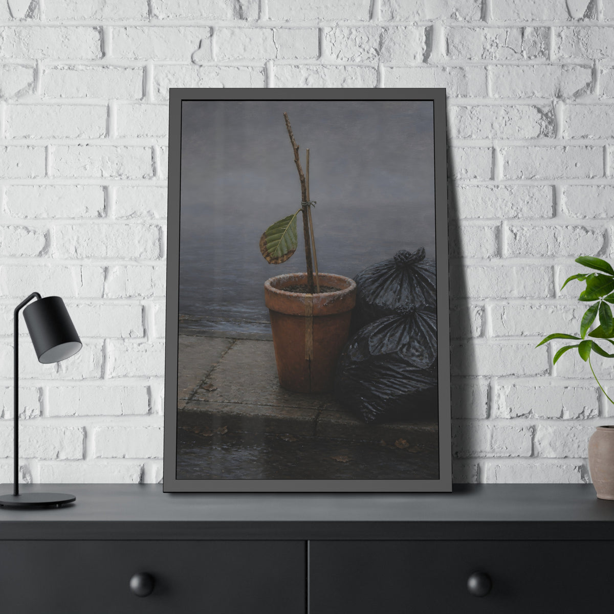 A potted dead twig with one leaf beside black garbage bags on a wet curb. The Curb on Tuesday by Garabaggio, framed fine art print hanging in a room.
