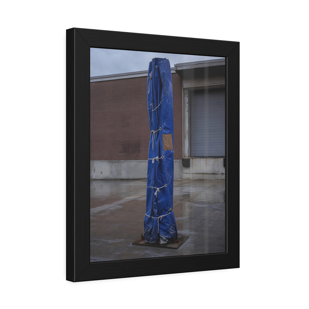 A tall blue tarp bundle roped upright on a wet lot by a loading dock. Folded Standard at the Loading Bay by Garabaggio, framed fine art print hanging in a room.