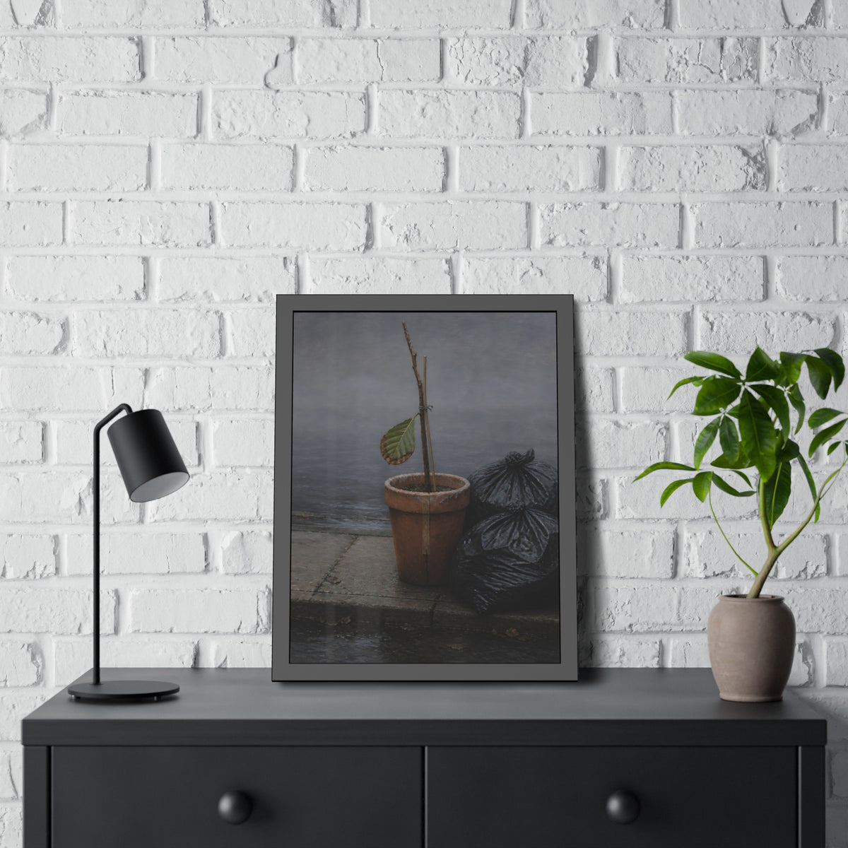 A potted dead twig with one leaf beside black garbage bags on a wet curb. The Curb on Tuesday by Garabaggio, framed fine art print hanging in a room.