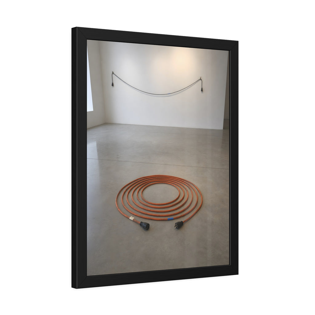 An orange extension cord coiled in a spiral on a bare gallery floor. The Floor Coil by Garabaggio, framed fine art print hanging in a room.