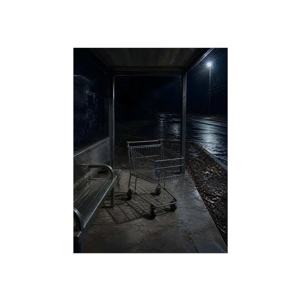 A shopping cart parked inside a bus shelter under a street lamp at night. The Rest at the Bus Shelter by Garabaggio, fine art print, 12″ x 16″.