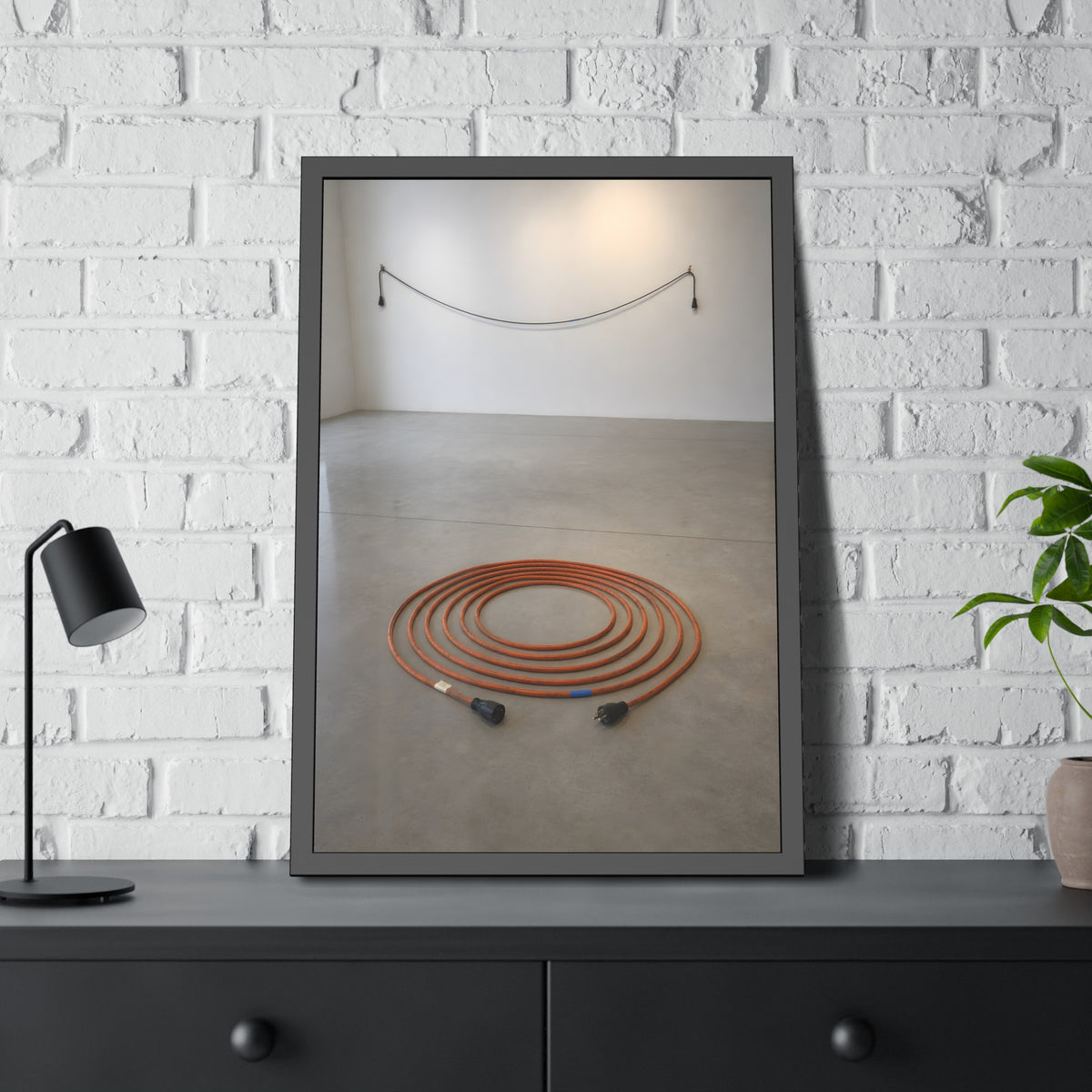 An orange extension cord coiled in a spiral on a bare gallery floor. The Floor Coil by Garabaggio, framed fine art print hanging in a room.