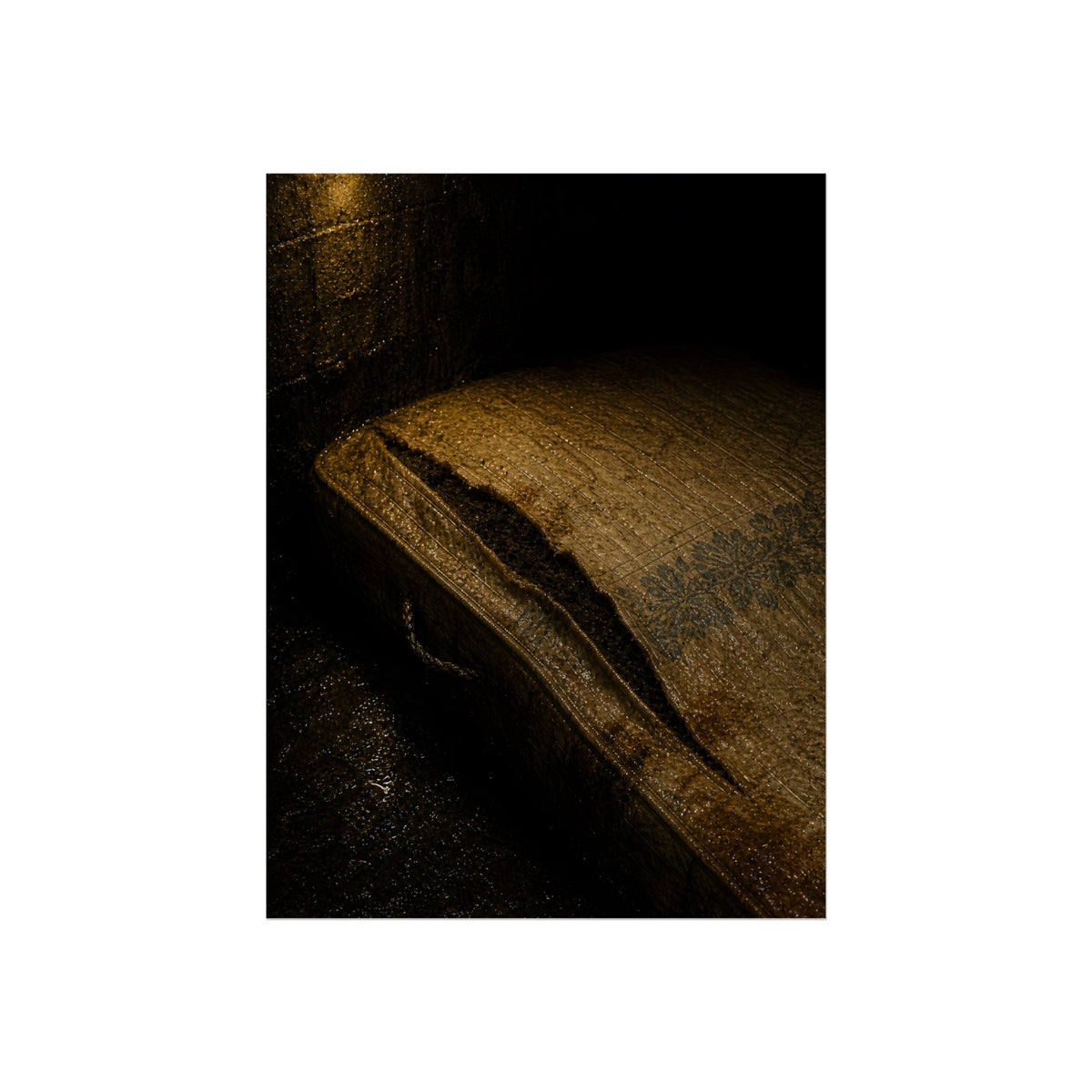 A mattress with its seam split open, lit warm against a dark wall. The Opening of the Ticking by Garabaggio, fine art print, 12″ x 16″.