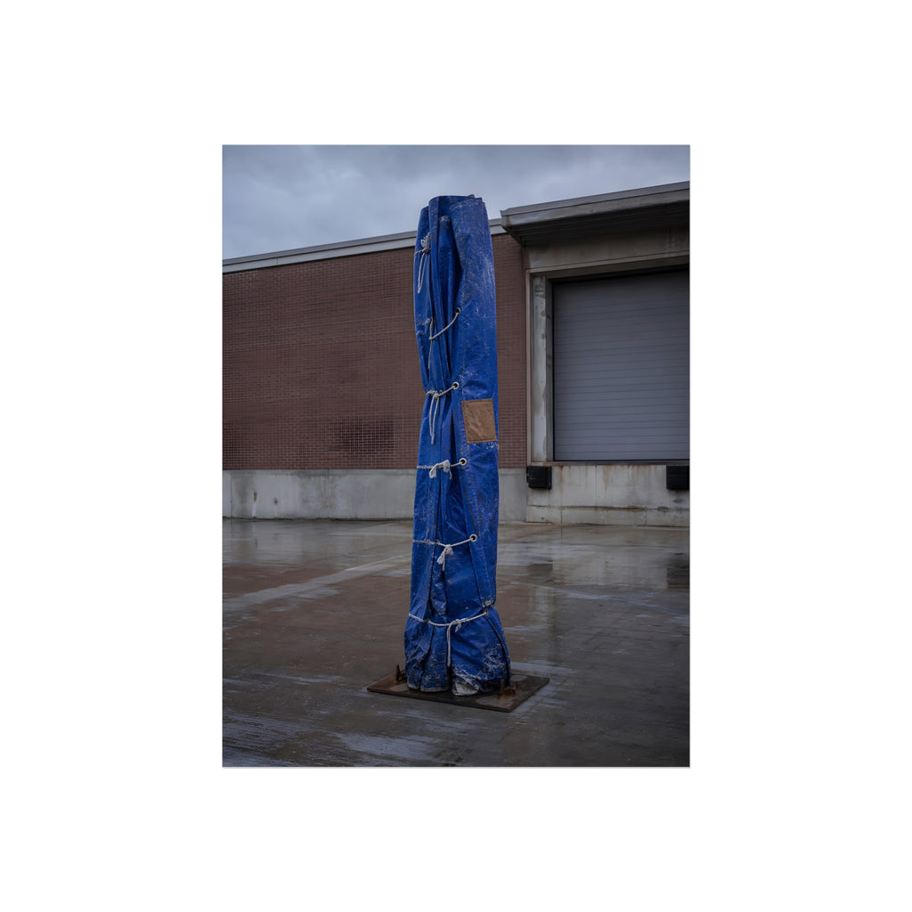 A tall blue tarp bundle roped upright on a wet lot by a loading dock. Folded Standard at the Loading Bay by Garabaggio, fine art print, 12″ x 16″.
