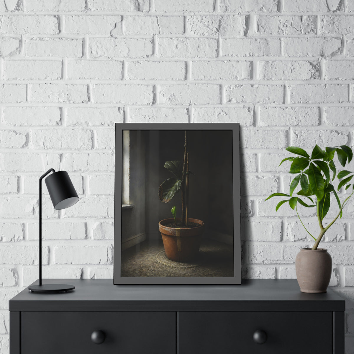A potted plant down to two curled leaves and one new sprout by a window. The Corner by the Window Again by Garabaggio, framed fine art print hanging in a room.