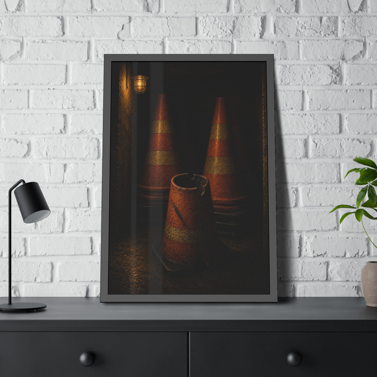 Stacked traffic cones behind a crushed one under a caged wall lamp. The Multiplication of Cones by Garabaggio, framed fine art print hanging in a room.