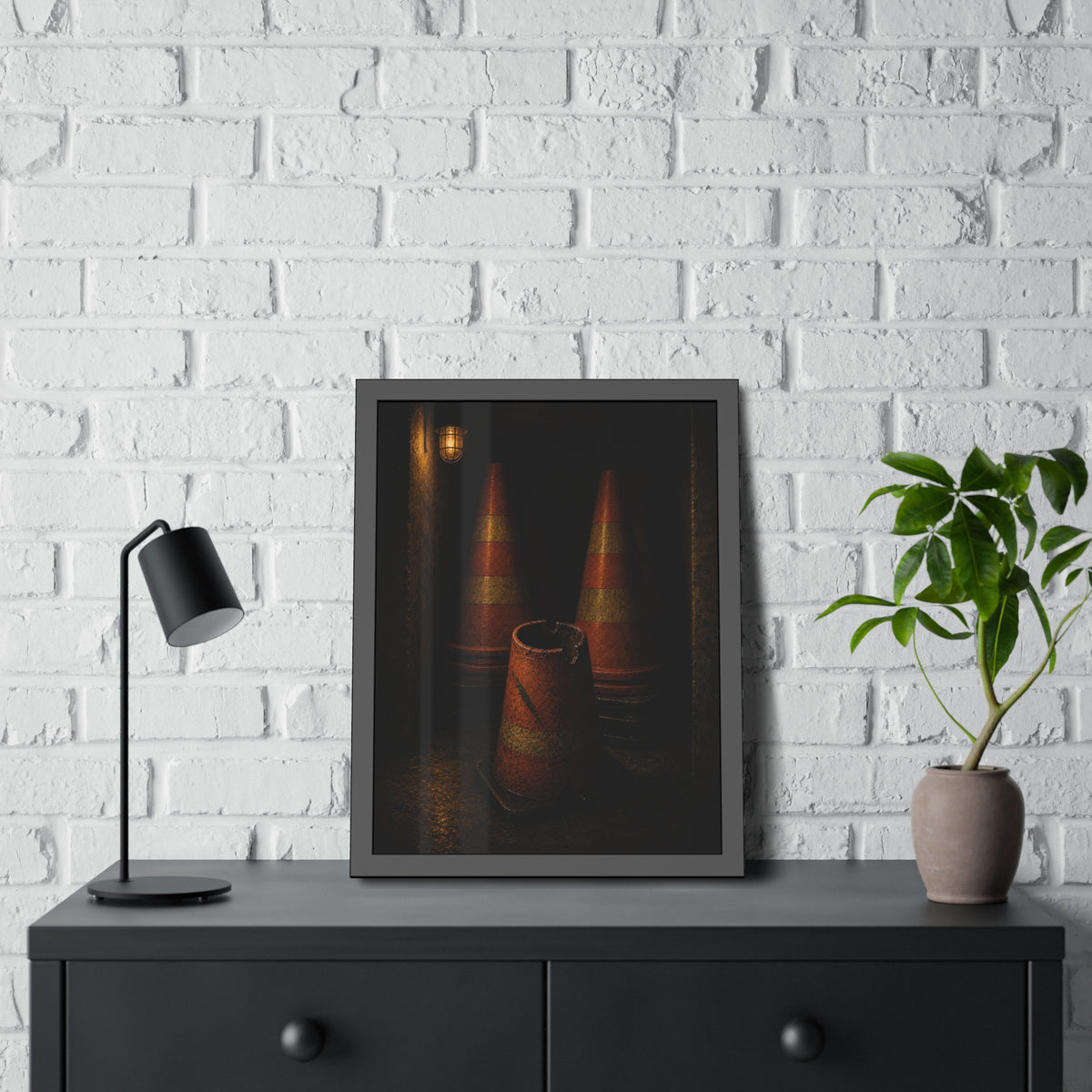 Stacked traffic cones behind a crushed one under a caged wall lamp. The Multiplication of Cones by Garabaggio, framed fine art print hanging in a room.