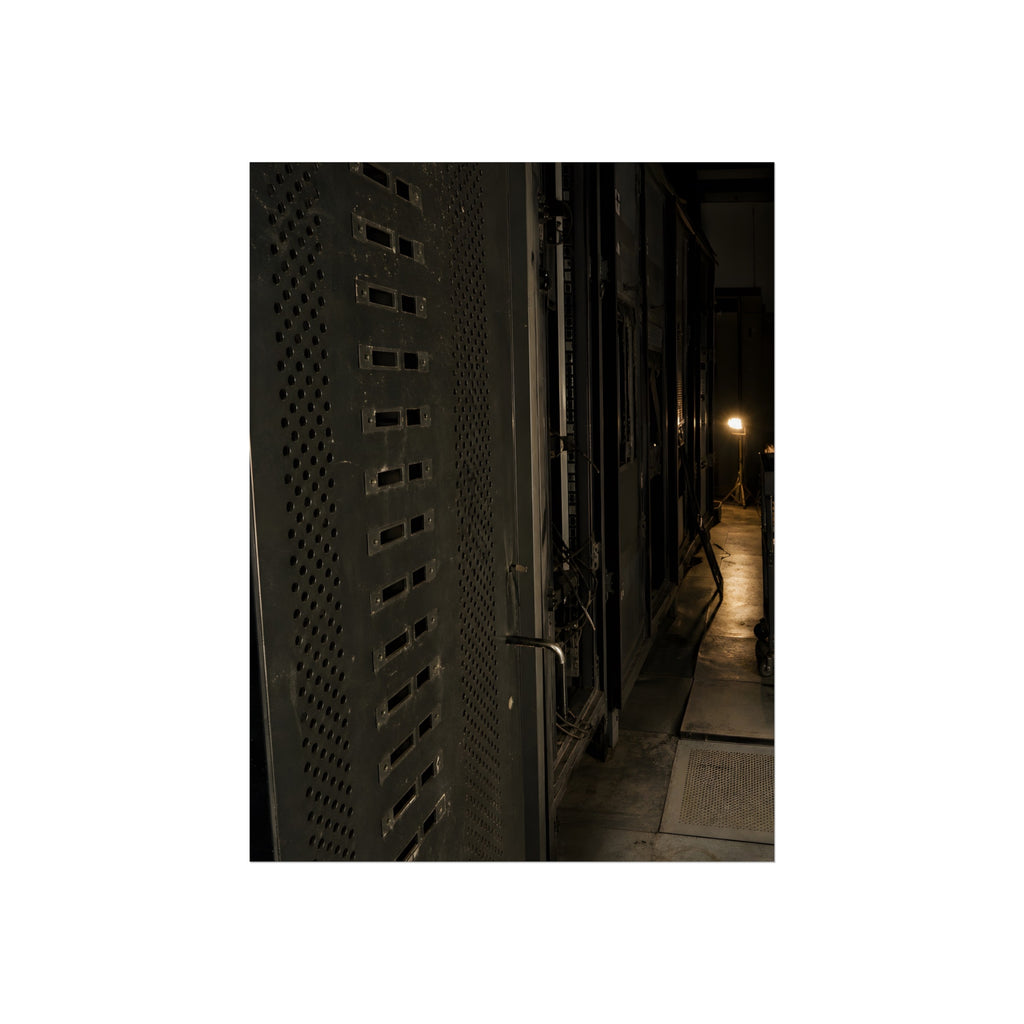 Rows of dark server cabinets with one work lamp glowing far down the aisle. Racks Without Light by Garabaggio, fine art print, 12″ x 16″.