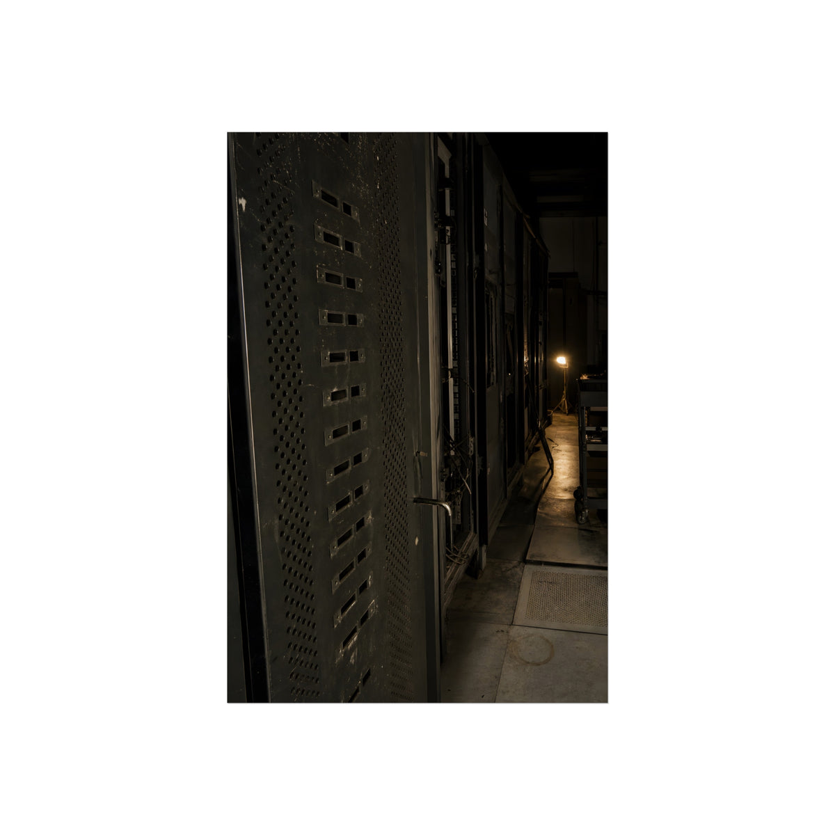 Rows of dark server cabinets with one work lamp glowing far down the aisle. Racks Without Light by Garabaggio, fine art print, 24″ x 36″.