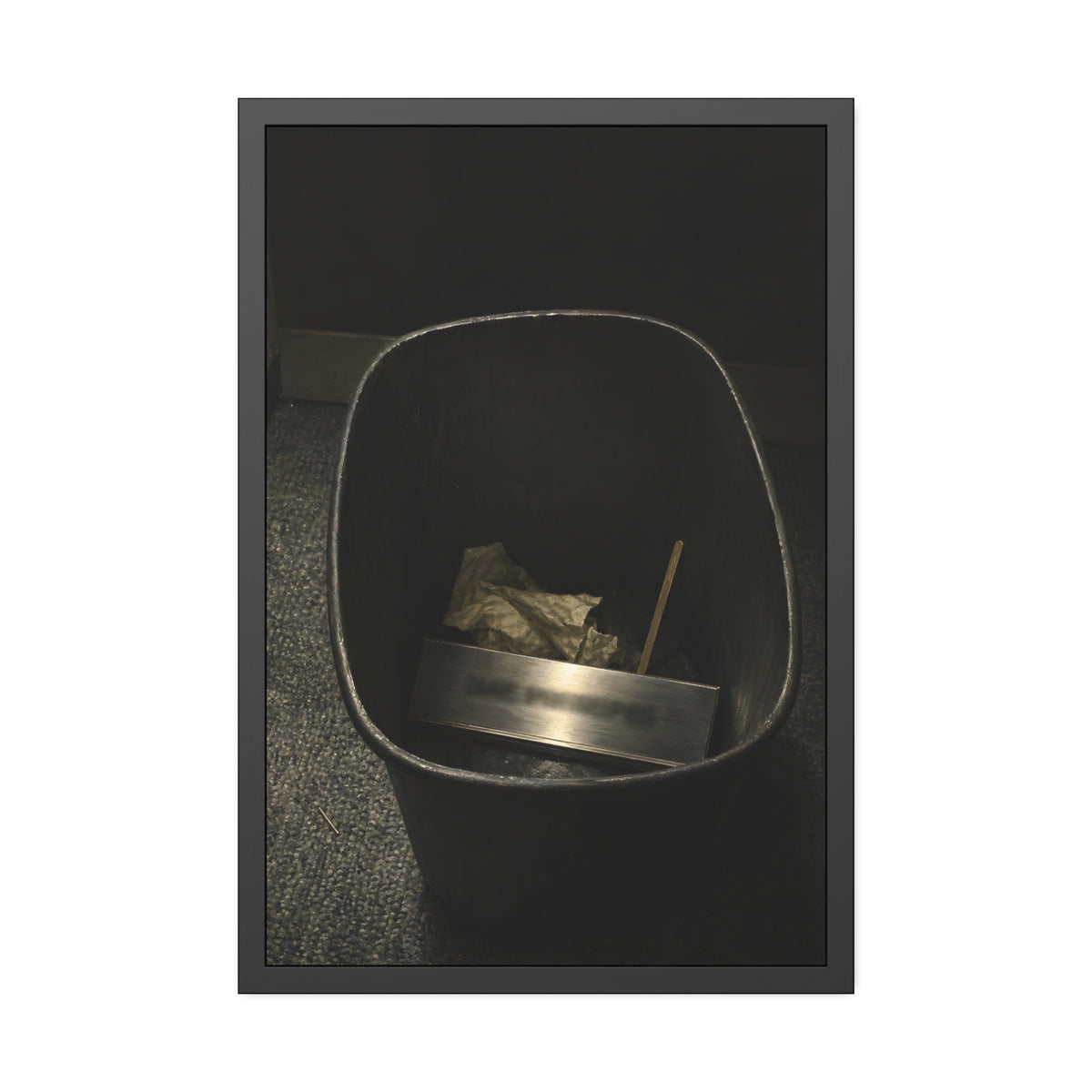 A metal nameplate lying in a black office waste bin on grey carpet. The Nameplate in the Waste Bin by Garabaggio, framed fine art print, 24″ x 36″.