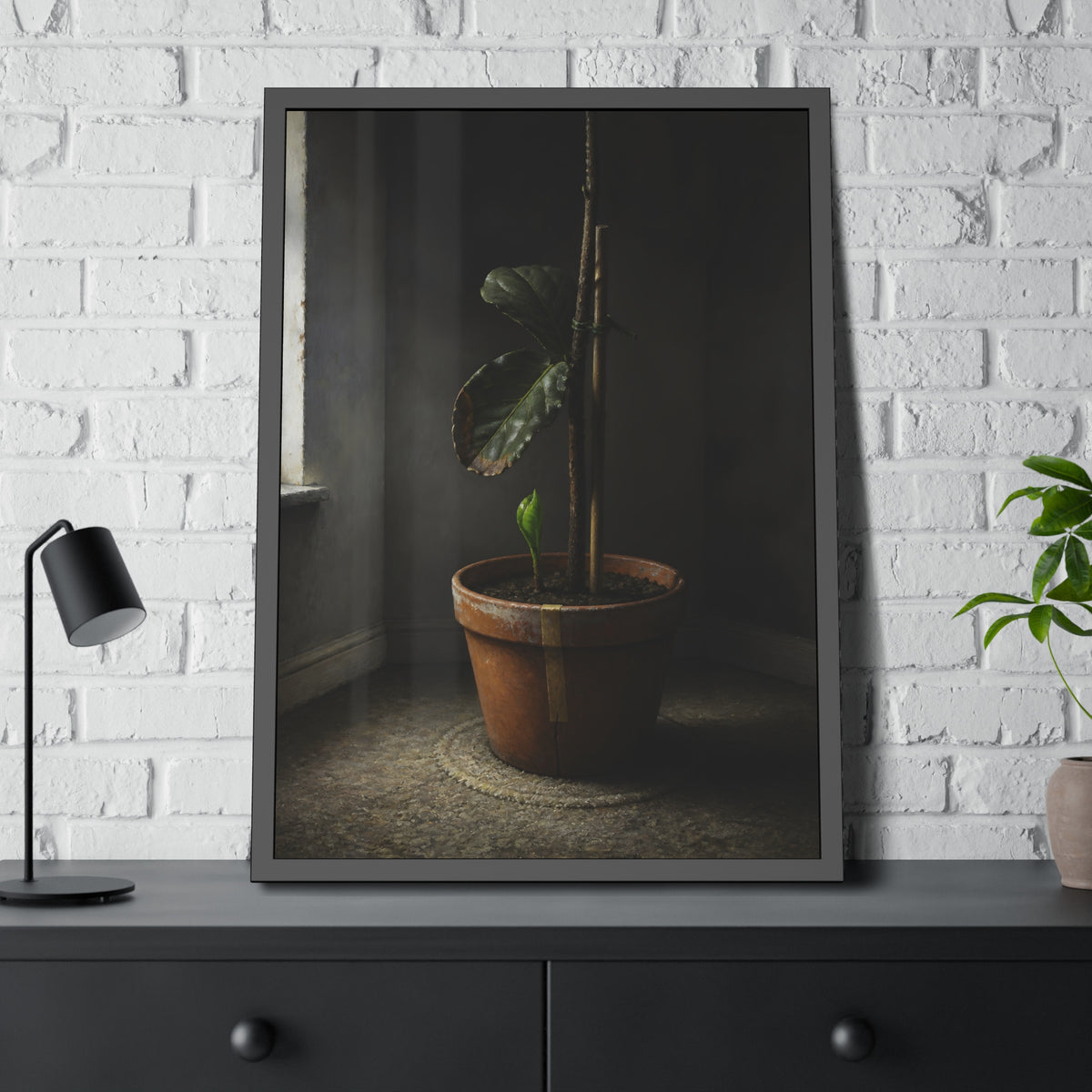 A potted plant down to two curled leaves and one new sprout by a window. The Corner by the Window Again by Garabaggio, framed fine art print hanging in a room.