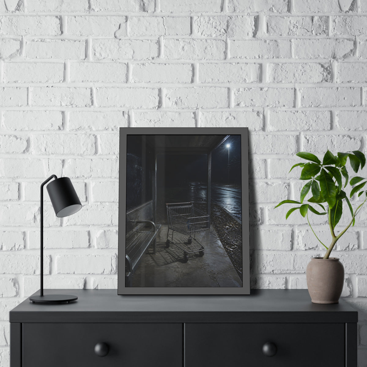 A shopping cart parked inside a bus shelter under a street lamp at night. The Rest at the Bus Shelter by Garabaggio, framed fine art print hanging in a room.