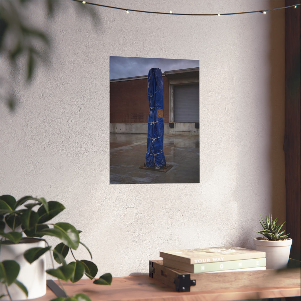 A tall blue tarp bundle roped upright on a wet lot by a loading dock. Folded Standard at the Loading Bay by Garabaggio, fine art print hanging in a room.