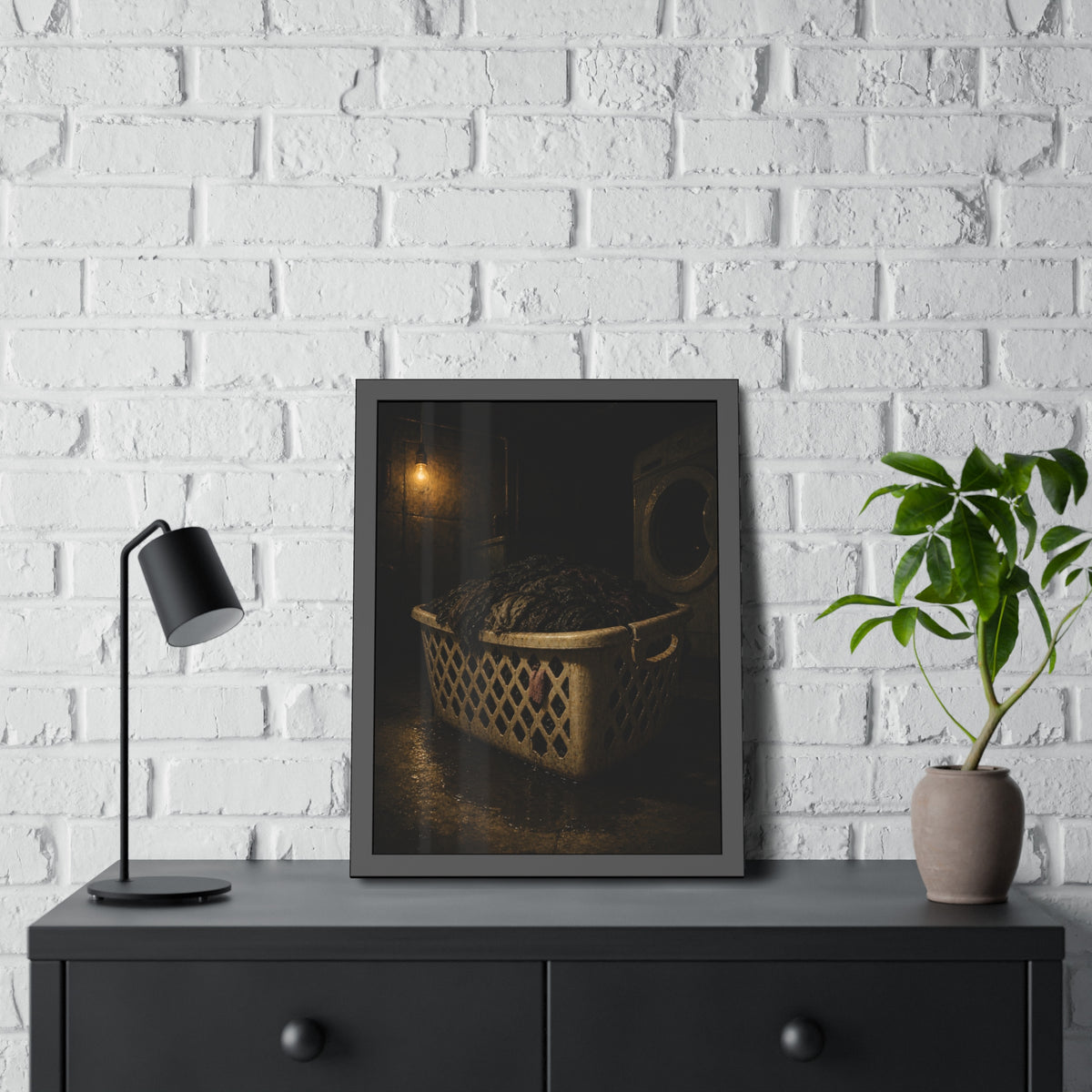 A laundry basket heaped with wet dark clothes under a bare bulb. The Burden of Wet Clothes by Garabaggio, framed fine art print hanging in a room.