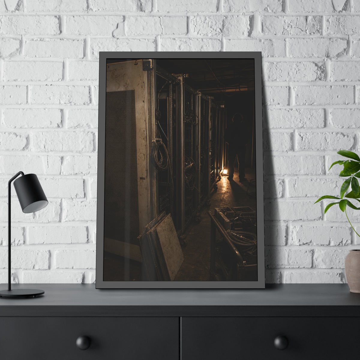 Open server racks in a dark room, a work lamp and a figure at the end. Garabaggio at the End of the Row by Garabaggio, framed fine art print hanging in a room.