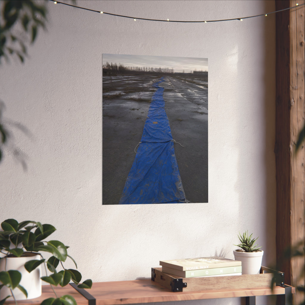 A blue tarp laid in a long strip down a wet cracked lot under grey sky. The Long Ground Procession by Garabaggio, fine art print hanging in a room.