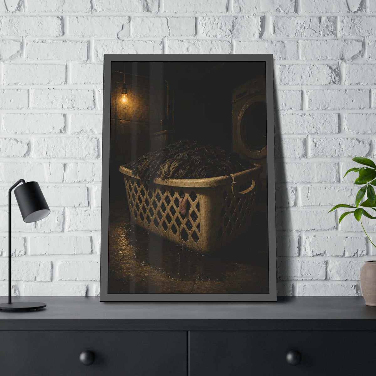 A laundry basket heaped with wet dark clothes under a bare bulb. The Burden of Wet Clothes by Garabaggio, framed fine art print hanging in a room.