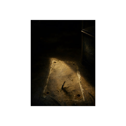 A worn floor patch with four bolt holes and a cut cable in a shaft of light. The Rectangle in the Floor by Garabaggio, fine art print, 18″ x 24″.