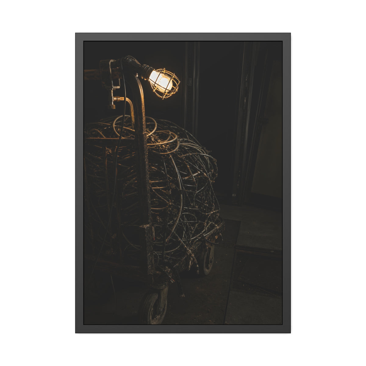 A tangled ball of cables on a hand cart under a caged work lamp. The Cable Harvest by Garabaggio, framed fine art print, 28″ x 40″.