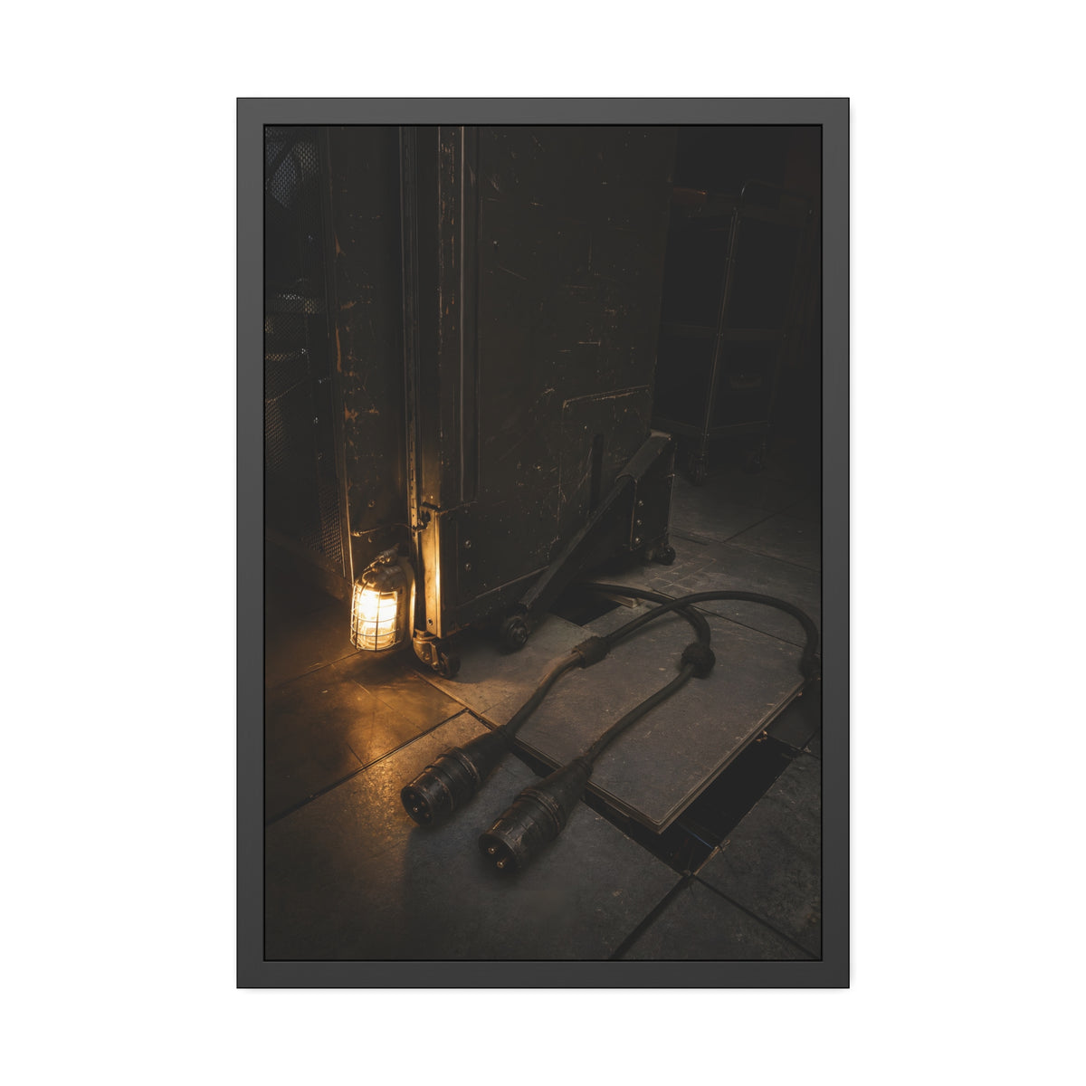 Two thick power cables unplugged on the floor beside a caged work lamp. The Redundant Power by Garabaggio, framed fine art print, 24″ x 36″.