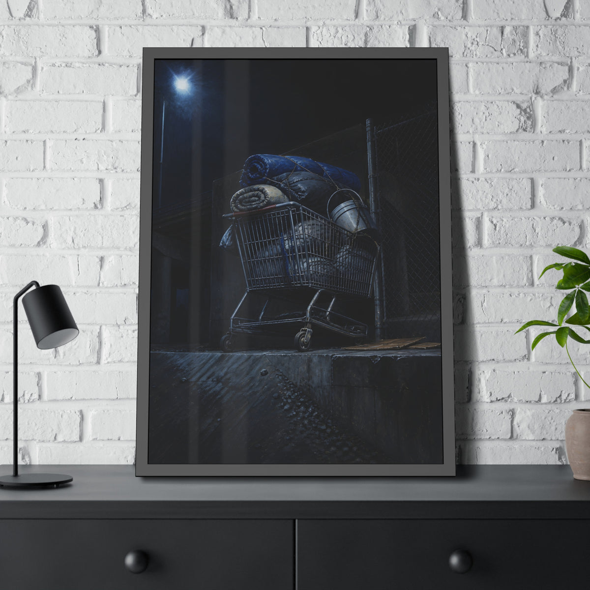 A shopping cart piled with bedding and buckets under a streetlamp. The Loading of Another Life by Garabaggio, framed fine art print hanging in a room.