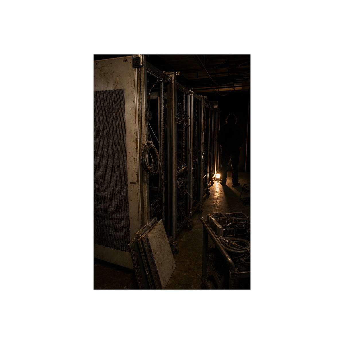 Open server racks in a dark room, a work lamp and a figure at the end. Garabaggio at the End of the Row by Garabaggio, fine art print, 24″ x 36″.