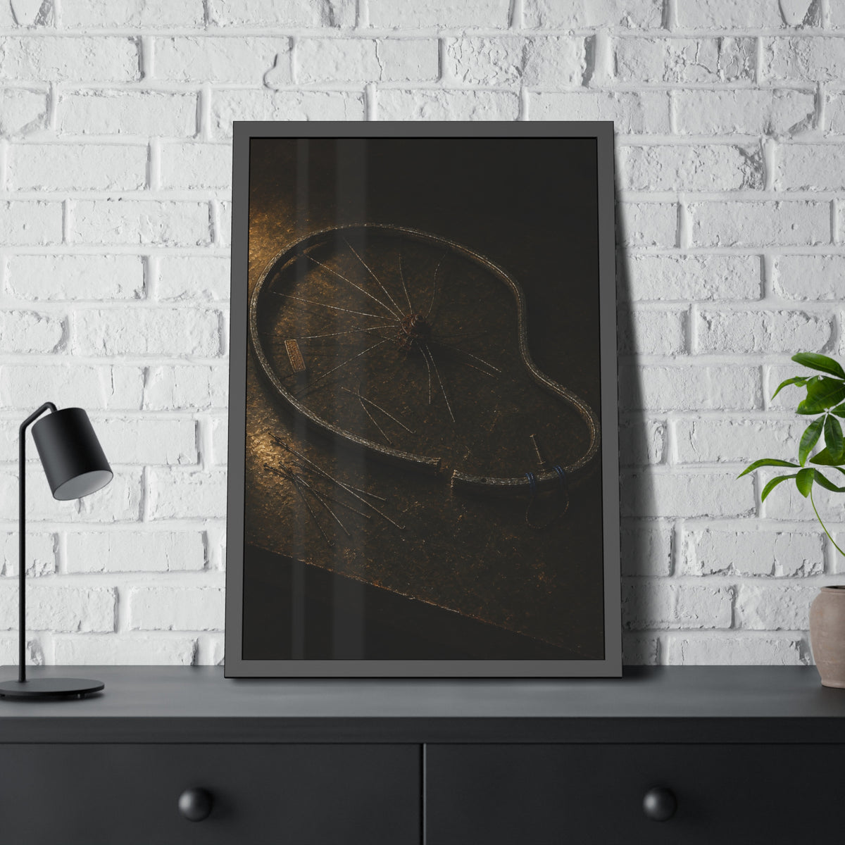 A bent bicycle wheel rim with loose spokes on a dark table in warm light. The Cutting of the Circle by Garabaggio, framed fine art print hanging in a room.