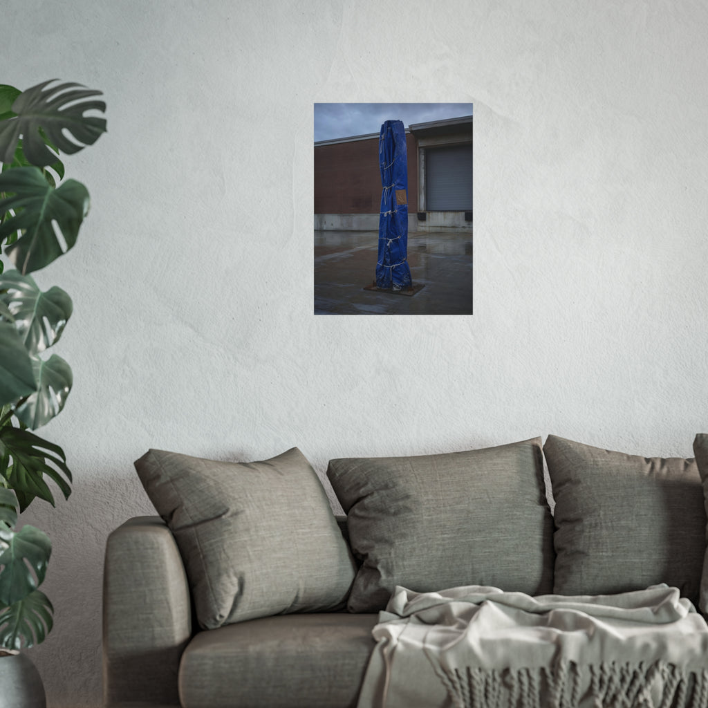 A tall blue tarp bundle roped upright on a wet lot by a loading dock. Folded Standard at the Loading Bay by Garabaggio, fine art print hanging in a room.