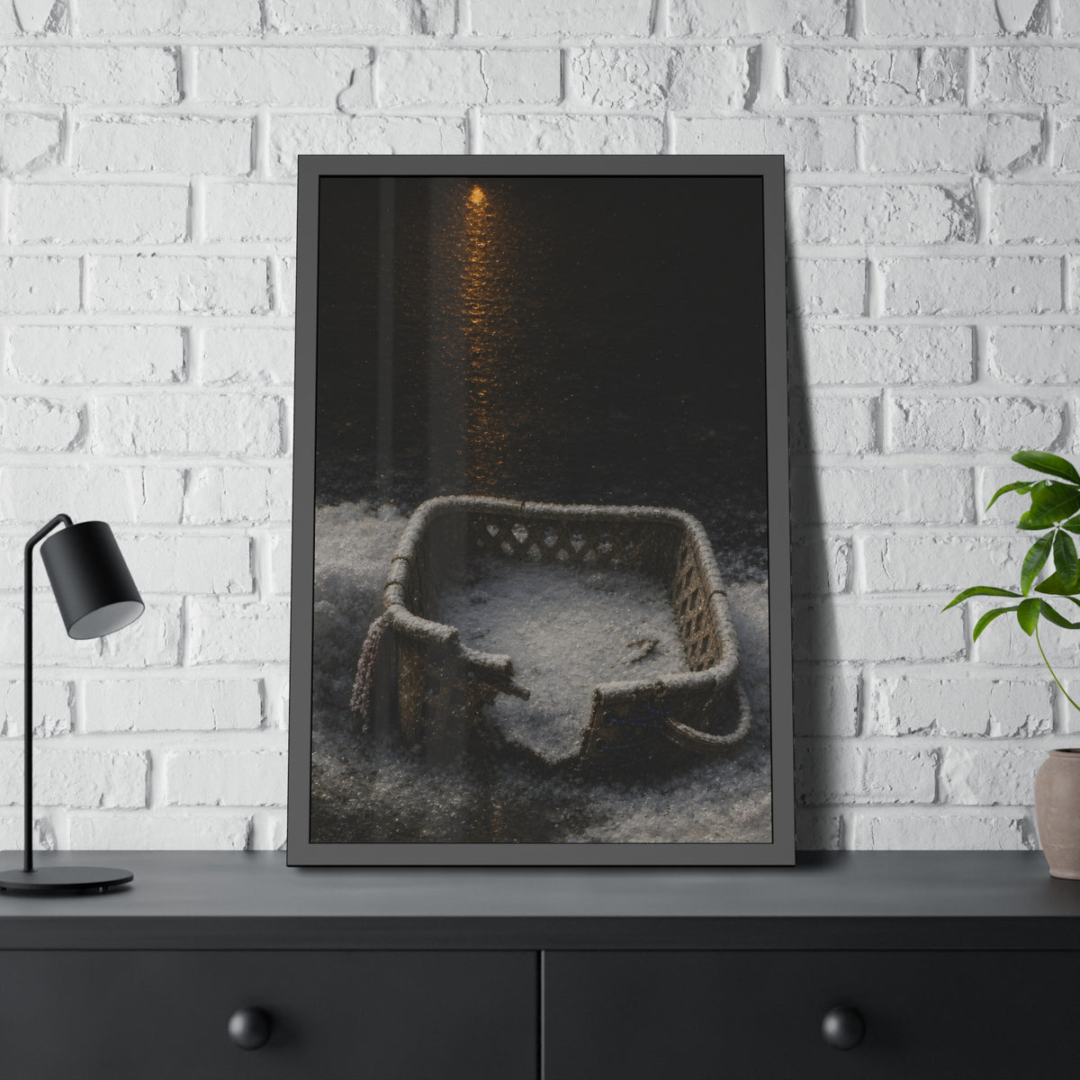 A cracked laundry basket half buried in snow under a distant lamp. The First Snow by Garabaggio, framed fine art print hanging in a room.