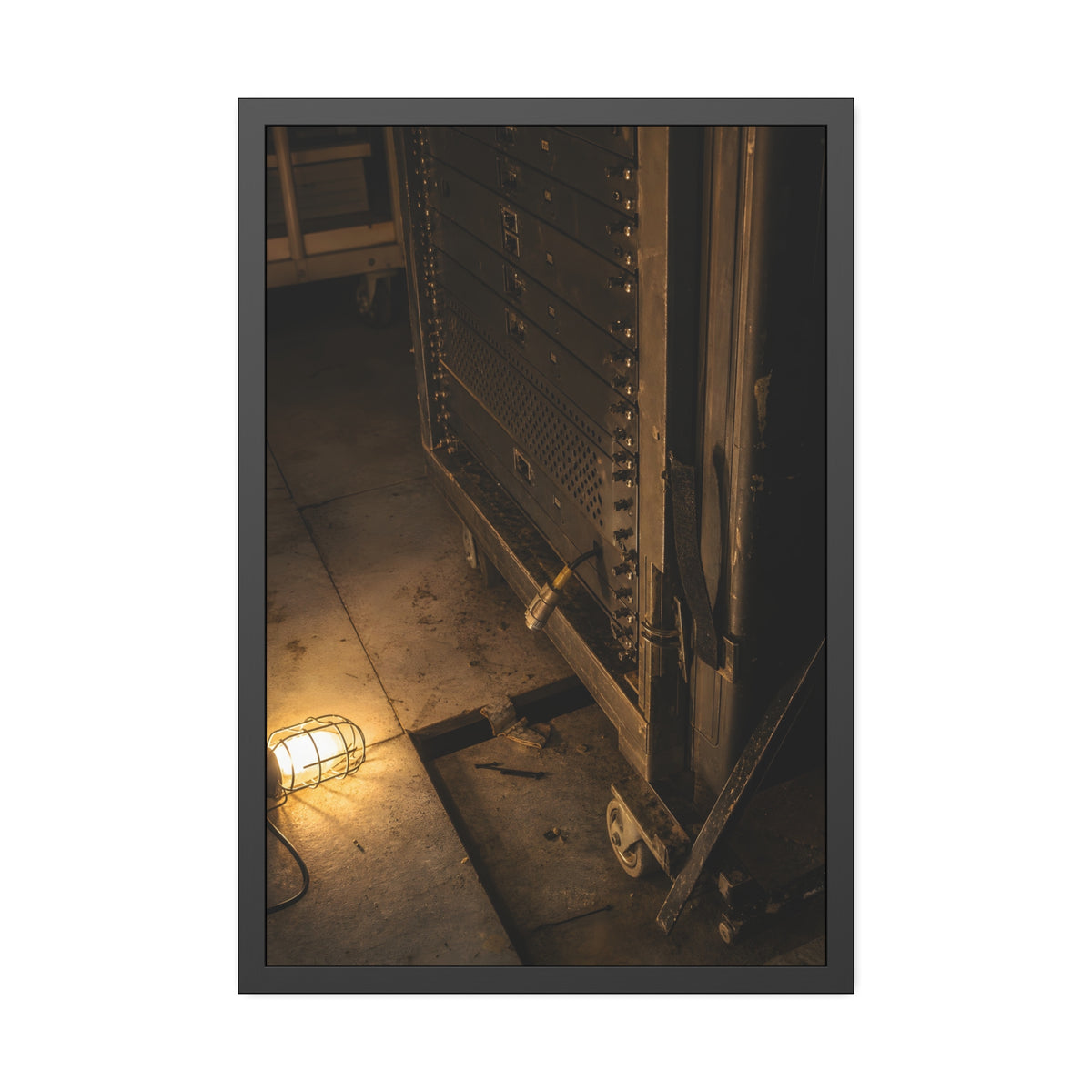 A server rack on a concrete floor beside a caged work light. The Silence at Cutover by Garabaggio, framed fine art print, 24″ x 36″.